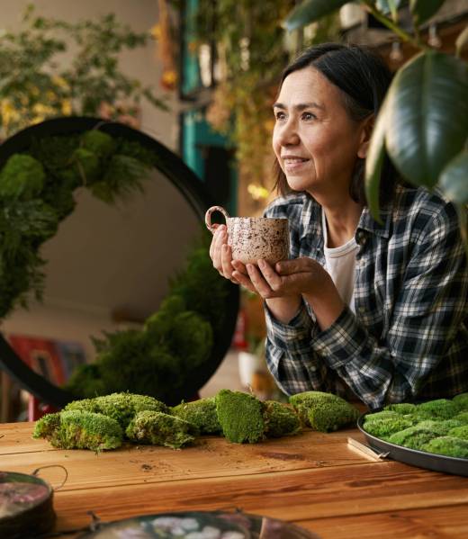 Designer holding cup of drink and resting after work Woman in plaid shirt sitting by table near creative artwork with green plants on mirror and drinking tea